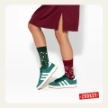 Dziecko w bordowej sukience i zielonych sneakersach, ma na sobie świąteczne skarpetki nie do pary. Zieloną z lampkami i bordową z reniferami i pierniczkami.