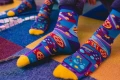 Stopy w kolorowych skarpetkach z motywem retro gier wideo, w żywych odcieniach niebieskiego, żółtego i fioletu, widoczne na kolorowej, geometrycznej wykładzinie.