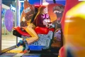 Osoba siedząca na czerwonym symulatorze motocykla w salonie gier, w kolorowych skarpetkach z motywem retro gier wideo, na tle maszyn z grafiką dinozaurów.