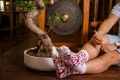 Mężczyzna siedzący na podłodze obok bonsai, w bordowo-białych skarpetkach z wzorem przypominającym plasterki ryby, trzymający w dłoni pałeczki.