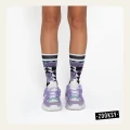 Nogi osoby stojącej w pastelowych sneakersach w odcieniach fioletu i mięty oraz artystycznych skarpetkach z geometrycznym wzorem w czerni, fiolecie i turkusie.